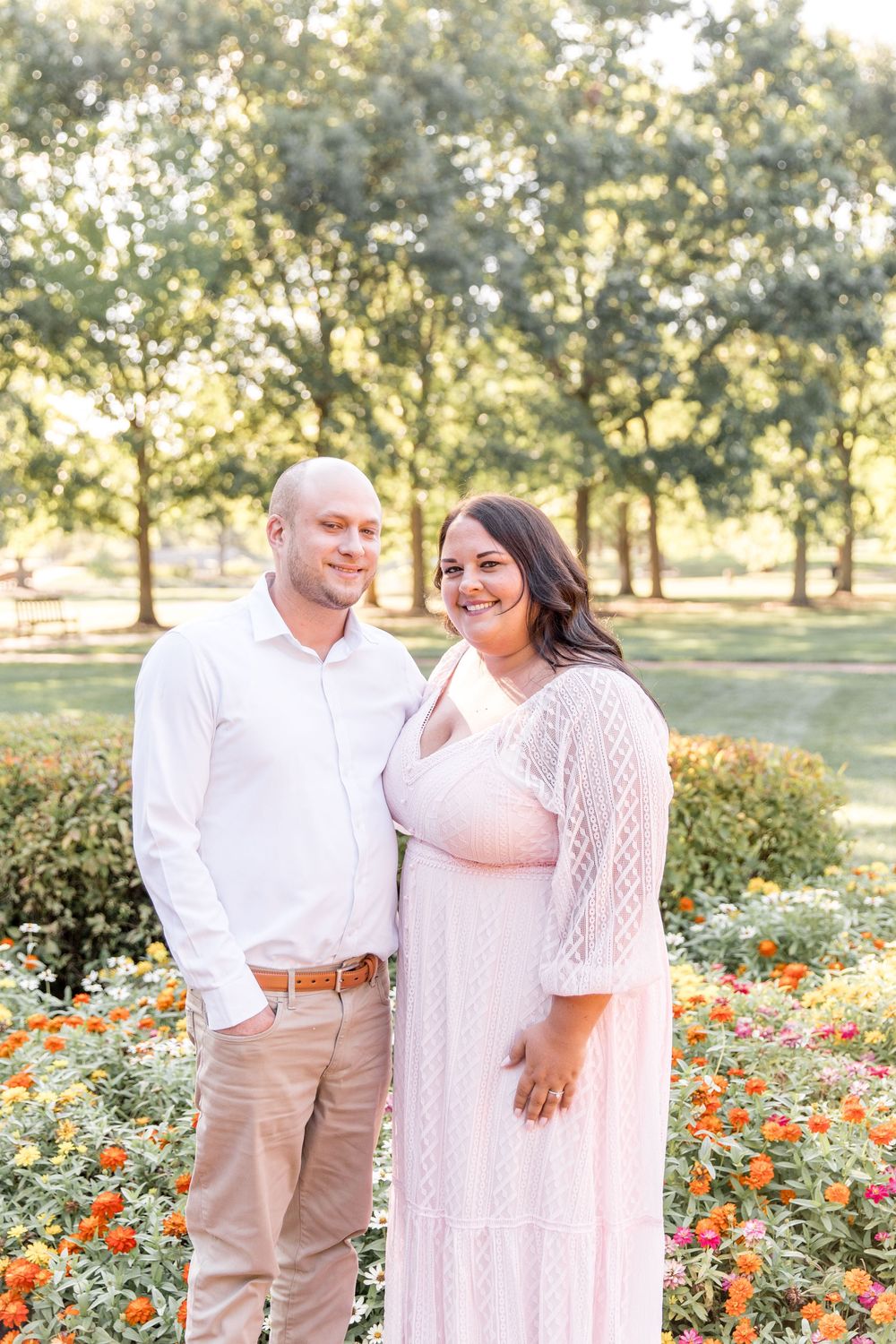 A couple stands together in a garden setting with blooming flowers and trees during golden hour lighting.