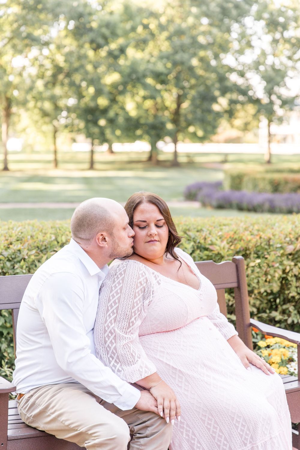 A couple in white and khaki shares an intimate moment while sitting on a wooden bench in a lush garden setting.