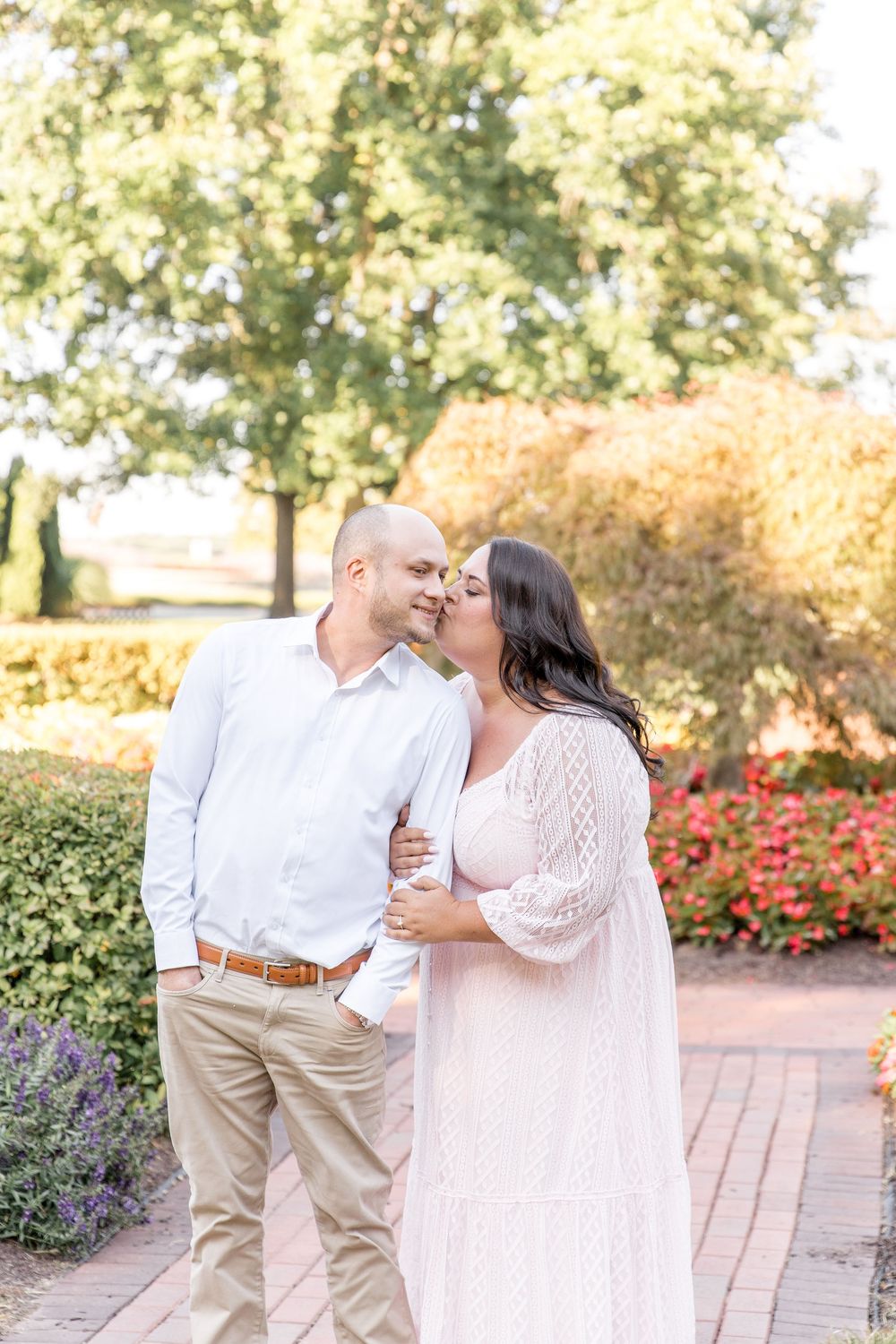 A couple shares an intimate moment together on a brick path surrounded by blooming gardens and trees on a sunny day.