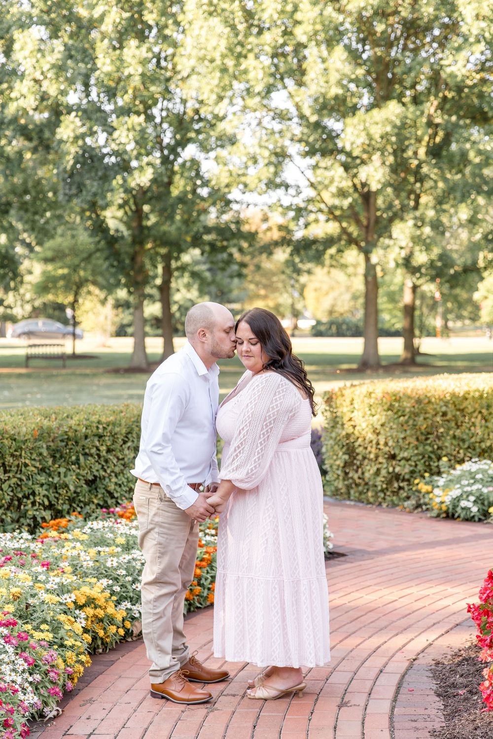 A couple shares tender moments along a curving brick pathway surrounded by vibrant flower gardens and trees at sunset.