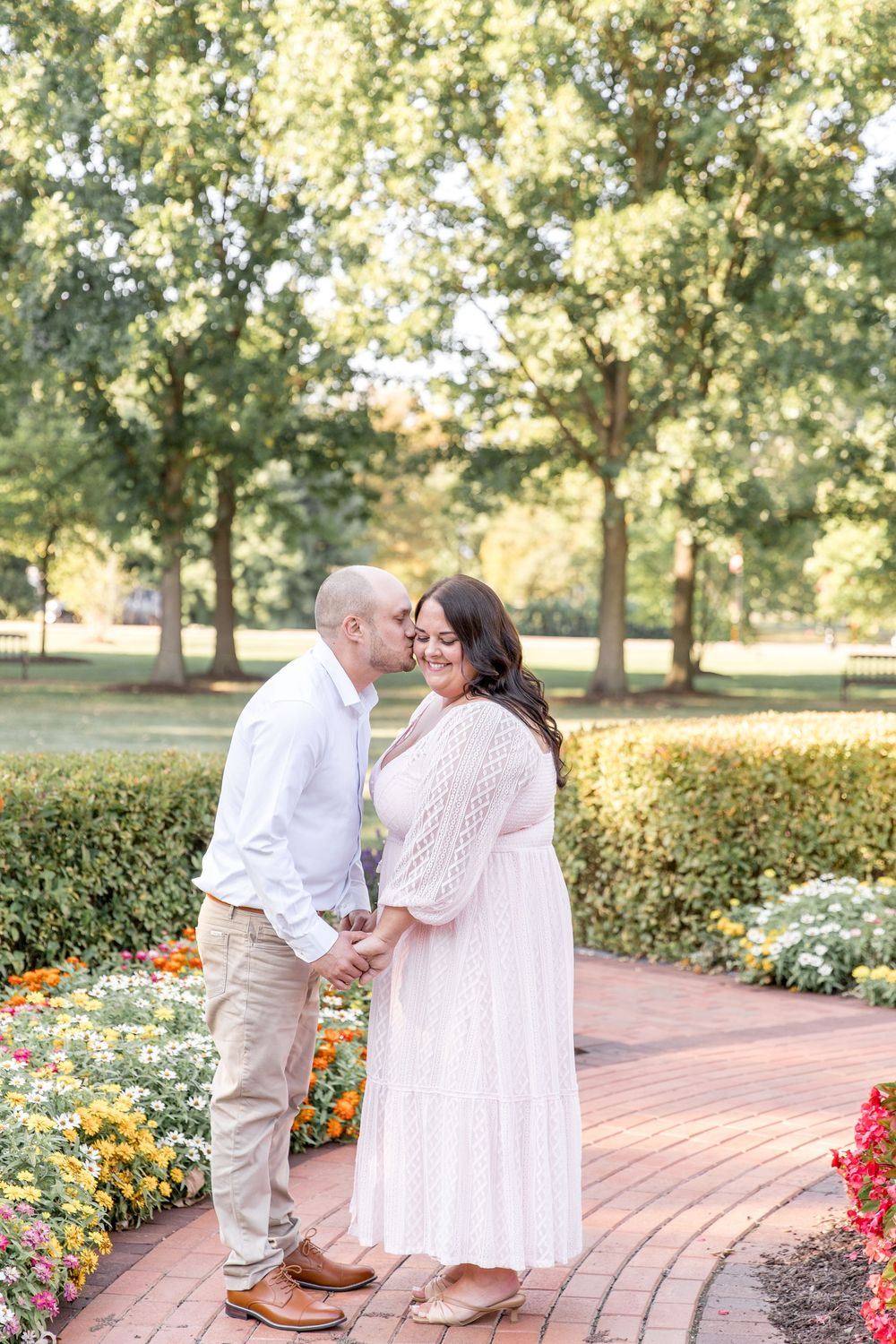 Couple sharing a romantic moment on a brick pathway surrounded by colorful flower gardens and trees in a park.