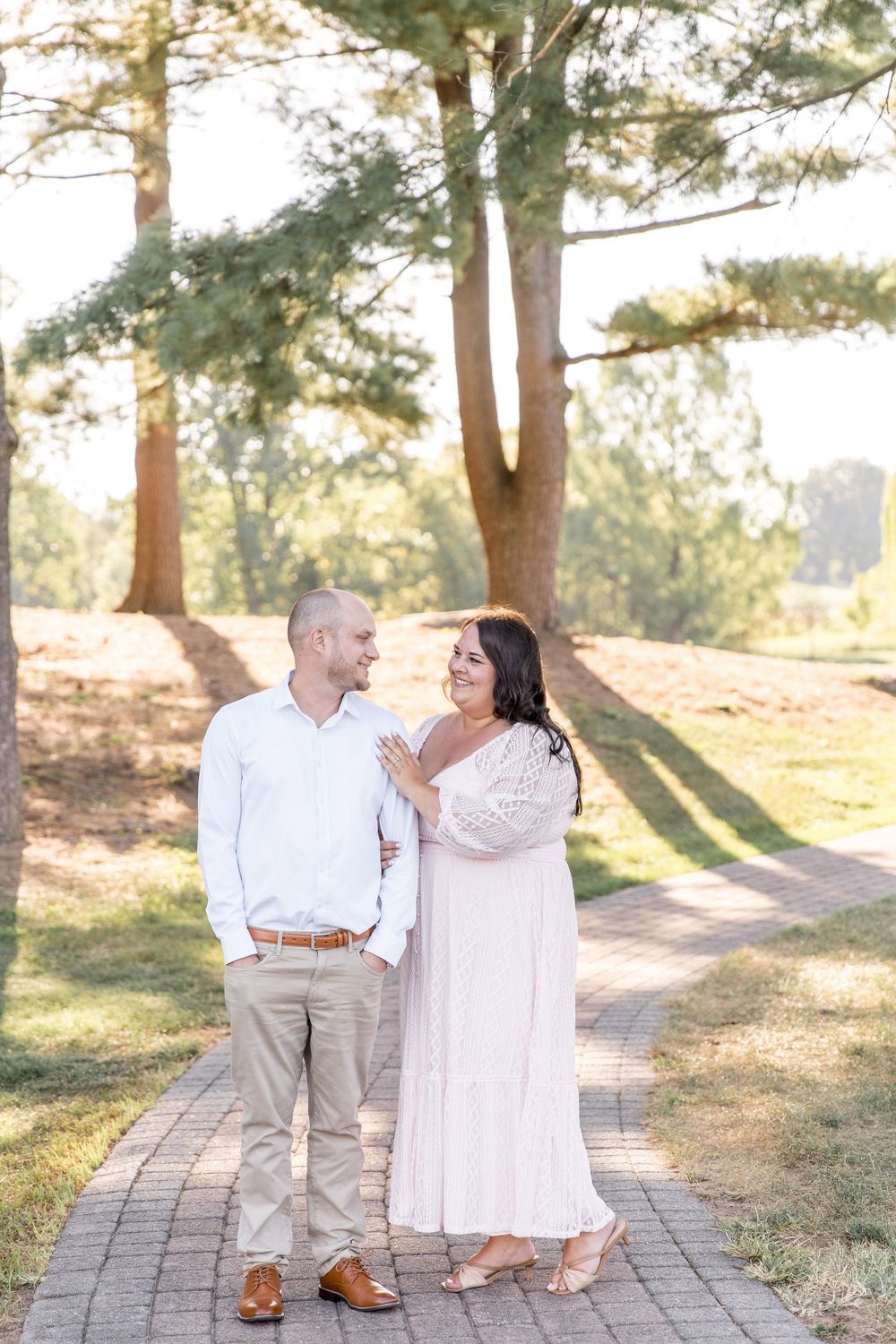 A couple shares a romantic moment while walking along a brick path surrounded by sunlit pine trees.