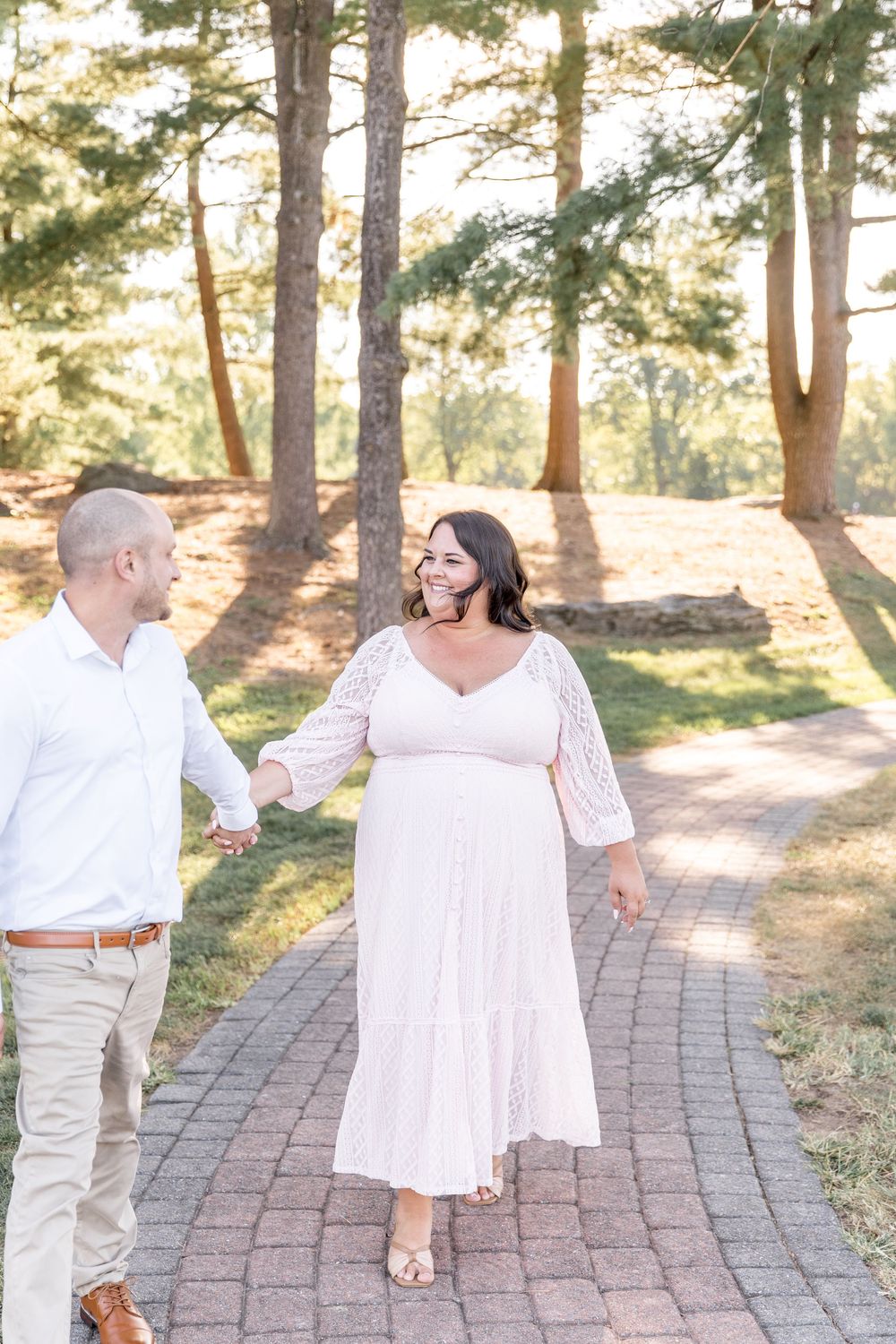 A couple in white outfits dances together on a brick path surrounded by sunlit pine trees during a photo session.