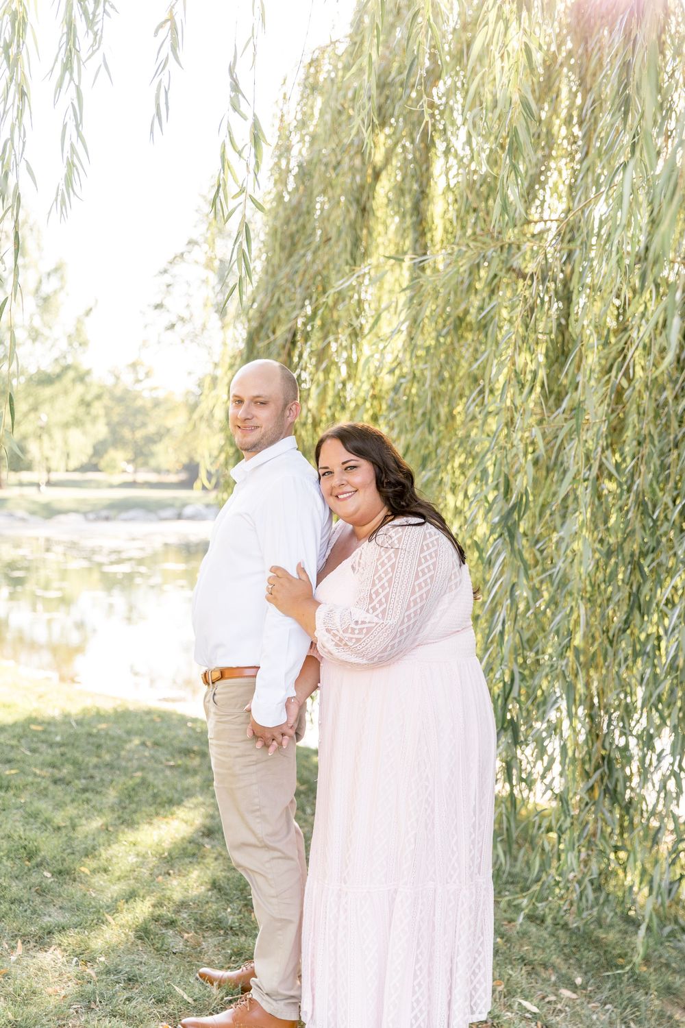 A couple in white attire shares romantic moments under a weeping willow tree in an outdoor setting.
