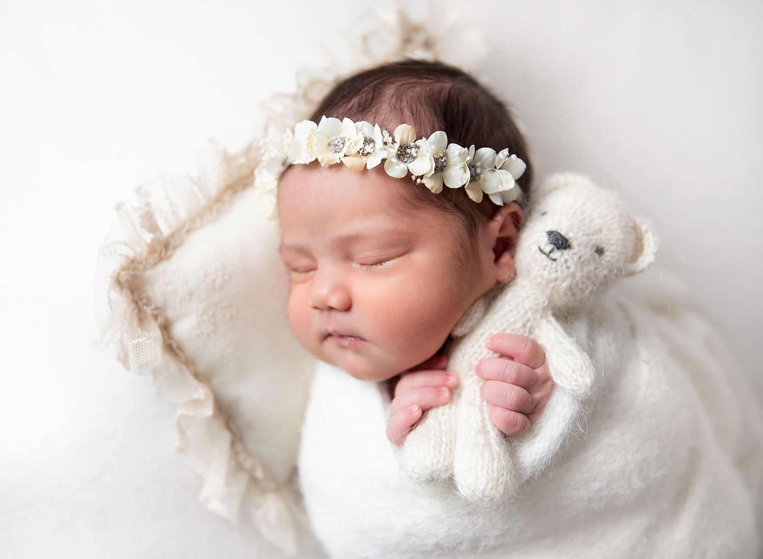 Peaceful sleeping newborn in white wrap holding small teddy bear with delicate floral crown on white backdrop. During a newborn session