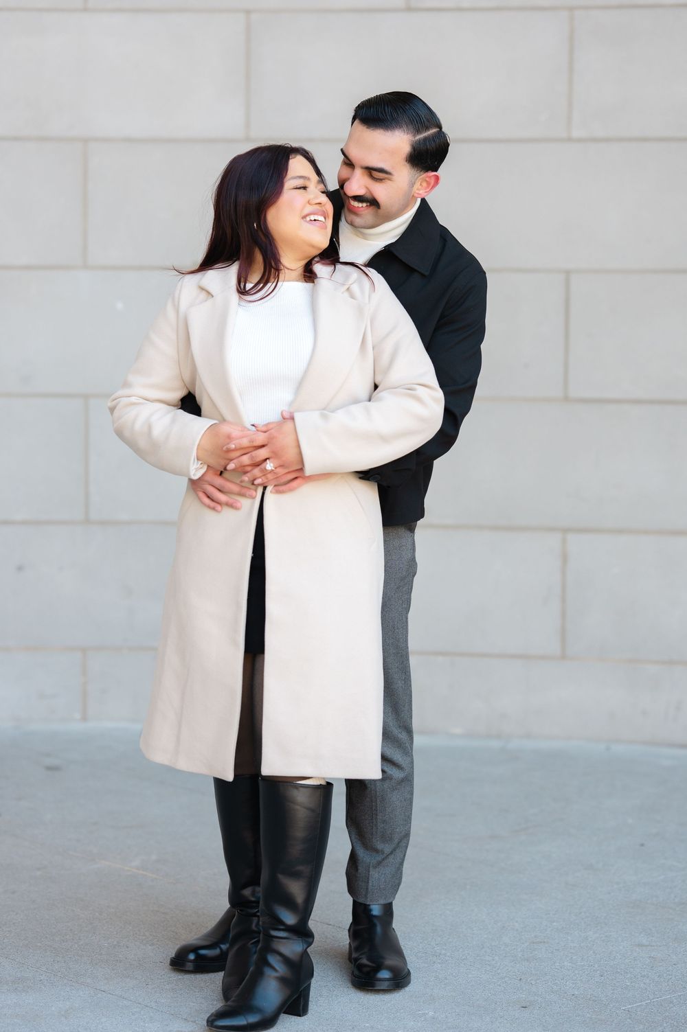 A romantic couple embraces while standing against a light colored wall, wearing coordinating winter coats.