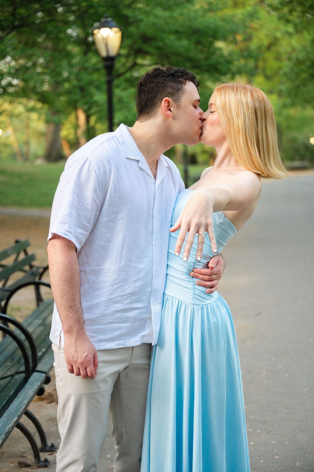 A couple shares a romantic kiss on a park path with a vintage lamp post and green foliage in the background.