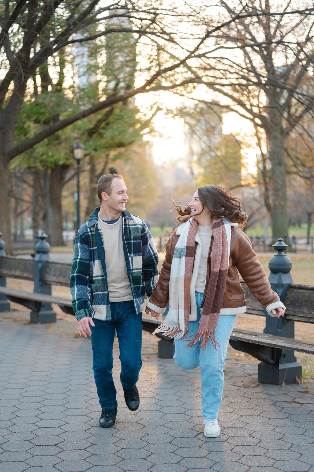 Two people strolling down a cobblestone path in a park on an autumn evening with warm sunlight filtering through trees.