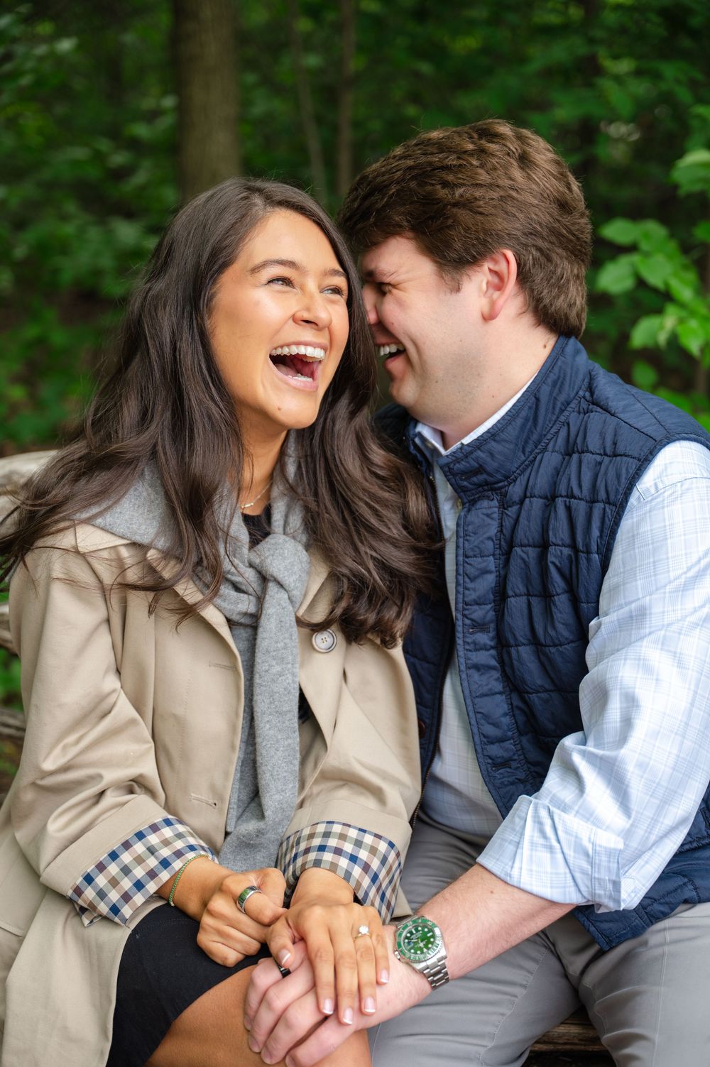 A couple joyfully laughs together while sitting outdoors, holding hands with a view of lush green trees in the background.