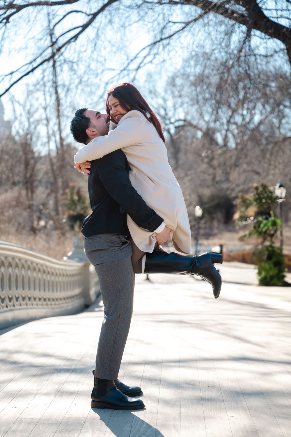 A couple shares a joyful embrace on a bridge path during winter, with bare trees in the background.