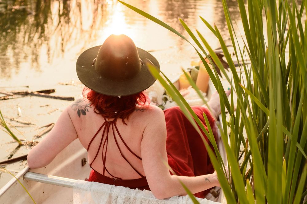 A person wearing a sun hat and pink shirt sits in a boat among reeds at sunset near a tranquil pond.