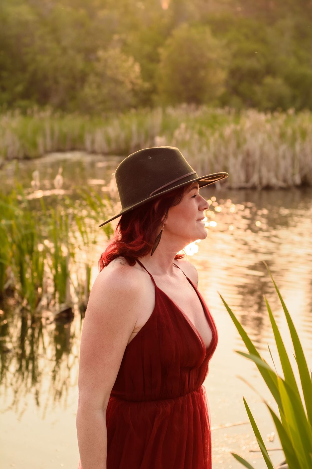 A figure in a burgundy dress and wide-brimmed hat stands near a serene pond at sunset with reeds in the background.