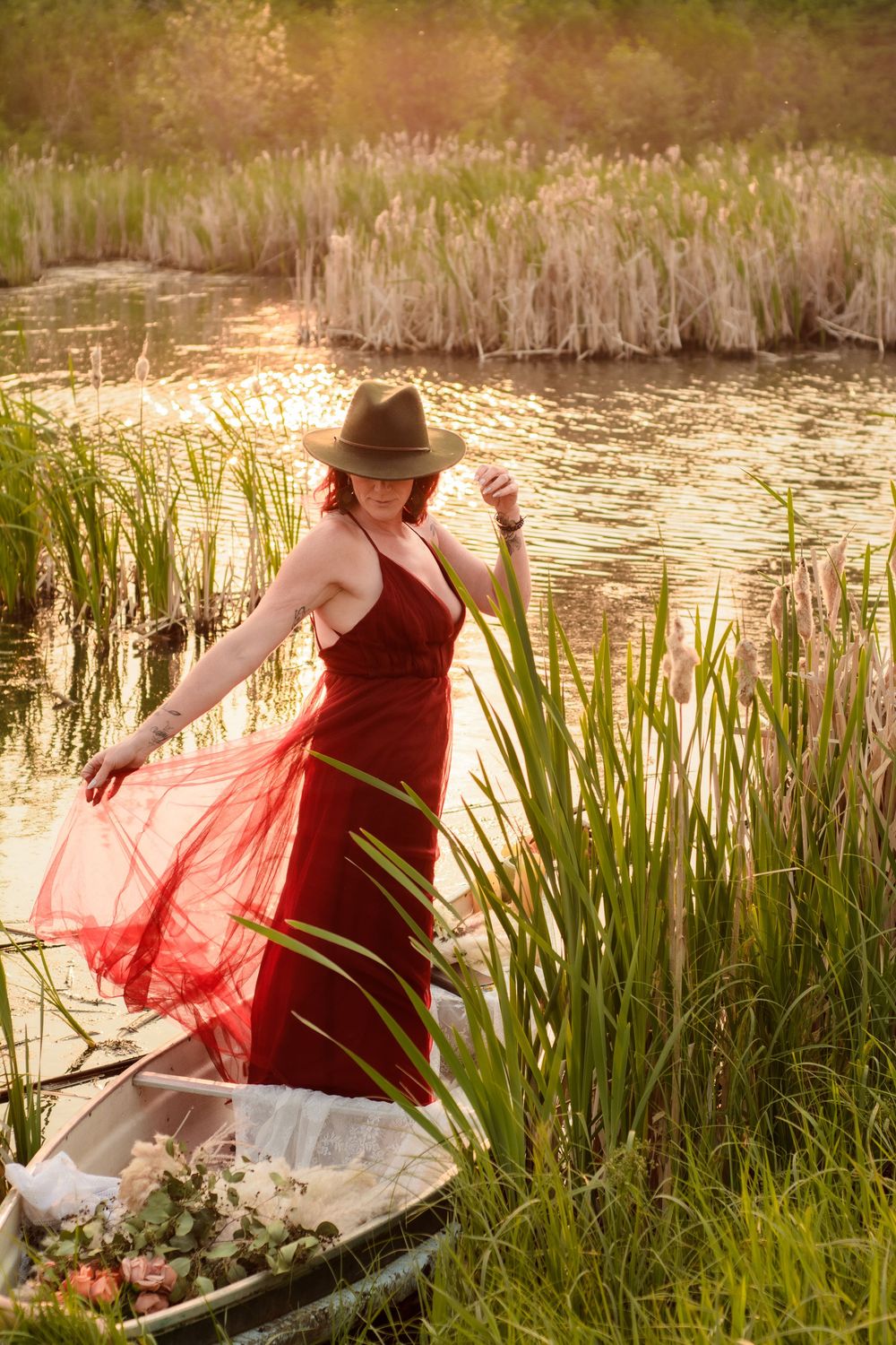 Woman in flowing red dress and hat stands gracefully among cattails by a pond at golden hour in summer.