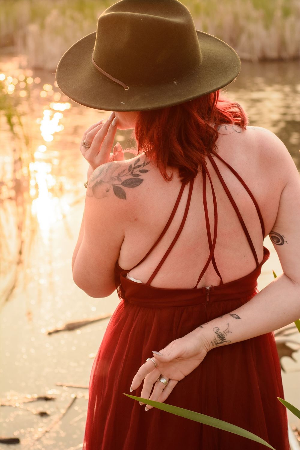 Person in red dress and brown hat poses by water at sunset with floral tattoo visible on shoulder.