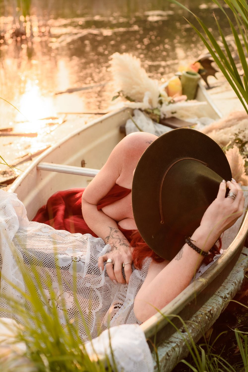 A person reclining in a vintage rowboat with a black hat and lace dress beside a tranquil pond at sunset.