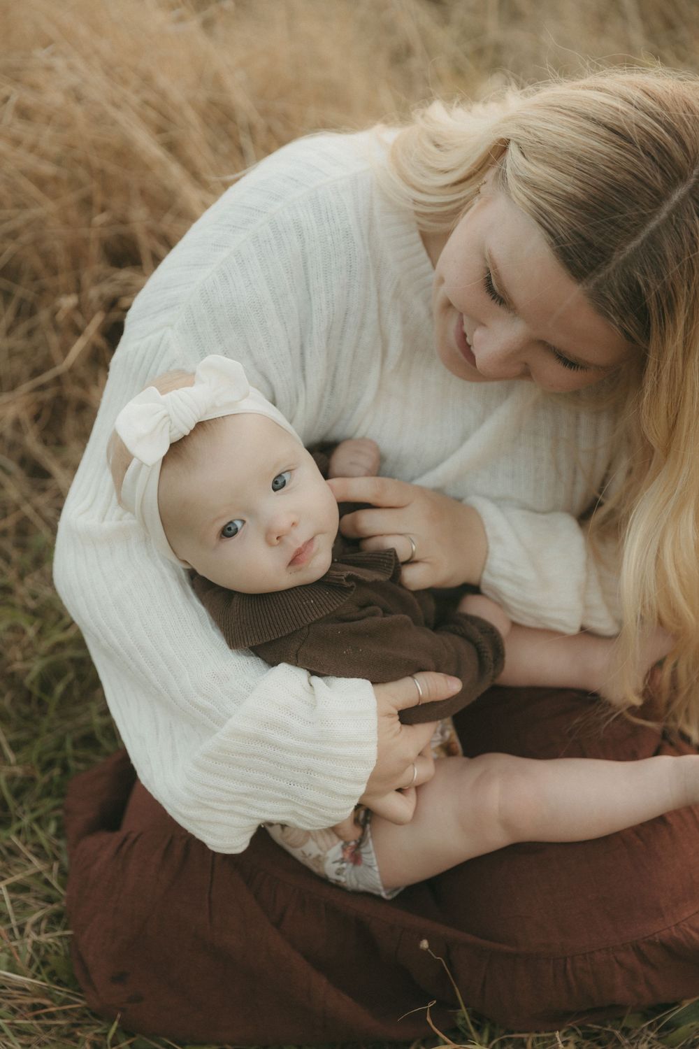 A mother in a white sweater holds her baby wearing brown clothing while sitting in a field of tall grass.