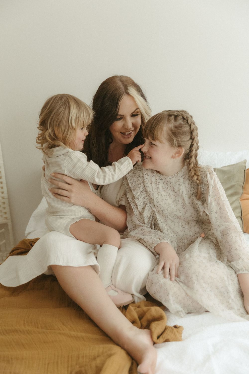 A parent and two children wearing white lace outfits share tender moments while sitting together on a mustard yellow bed.