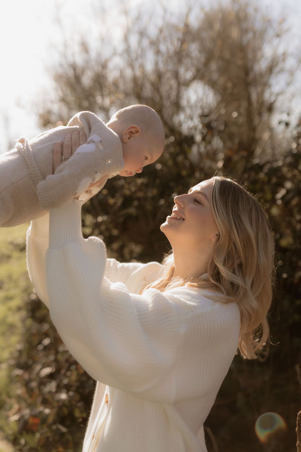 Joyful parent in white sweater lifts baby against sunlit background with trees on autumn day.