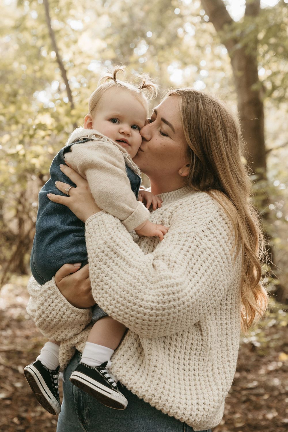 Tender moment outdoors as parent in chunky knit sweater holds and kisses baby wearing navy outfit in autumn setting.