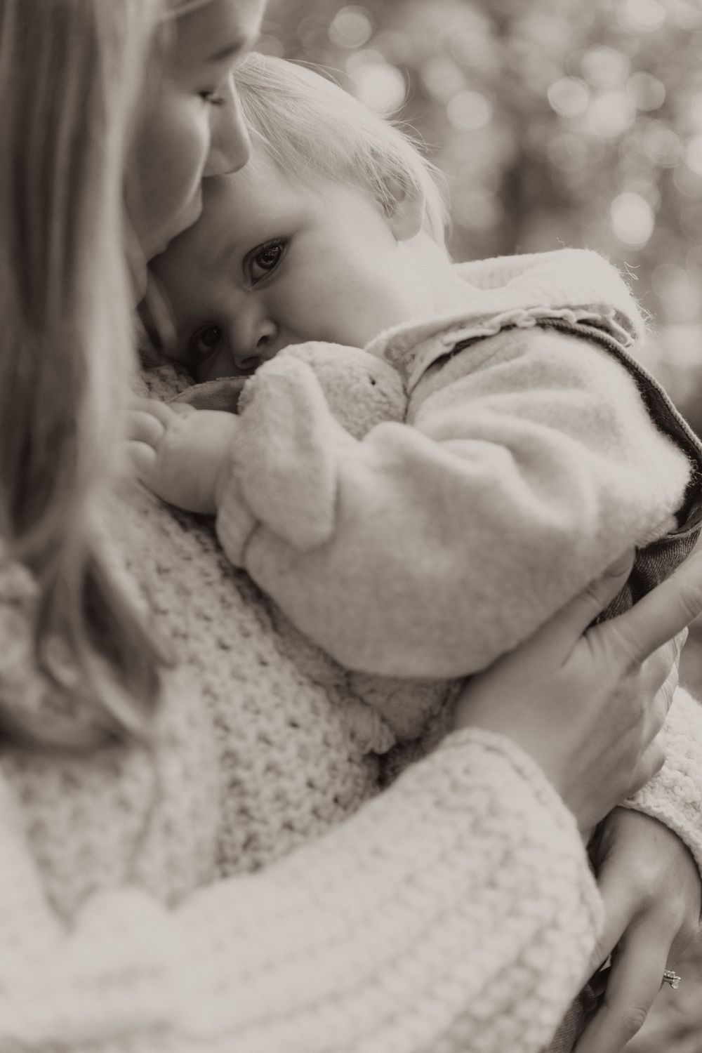 Black and white photo showing tender moment of parent holding young child in knit sweaters with soft bokeh background.