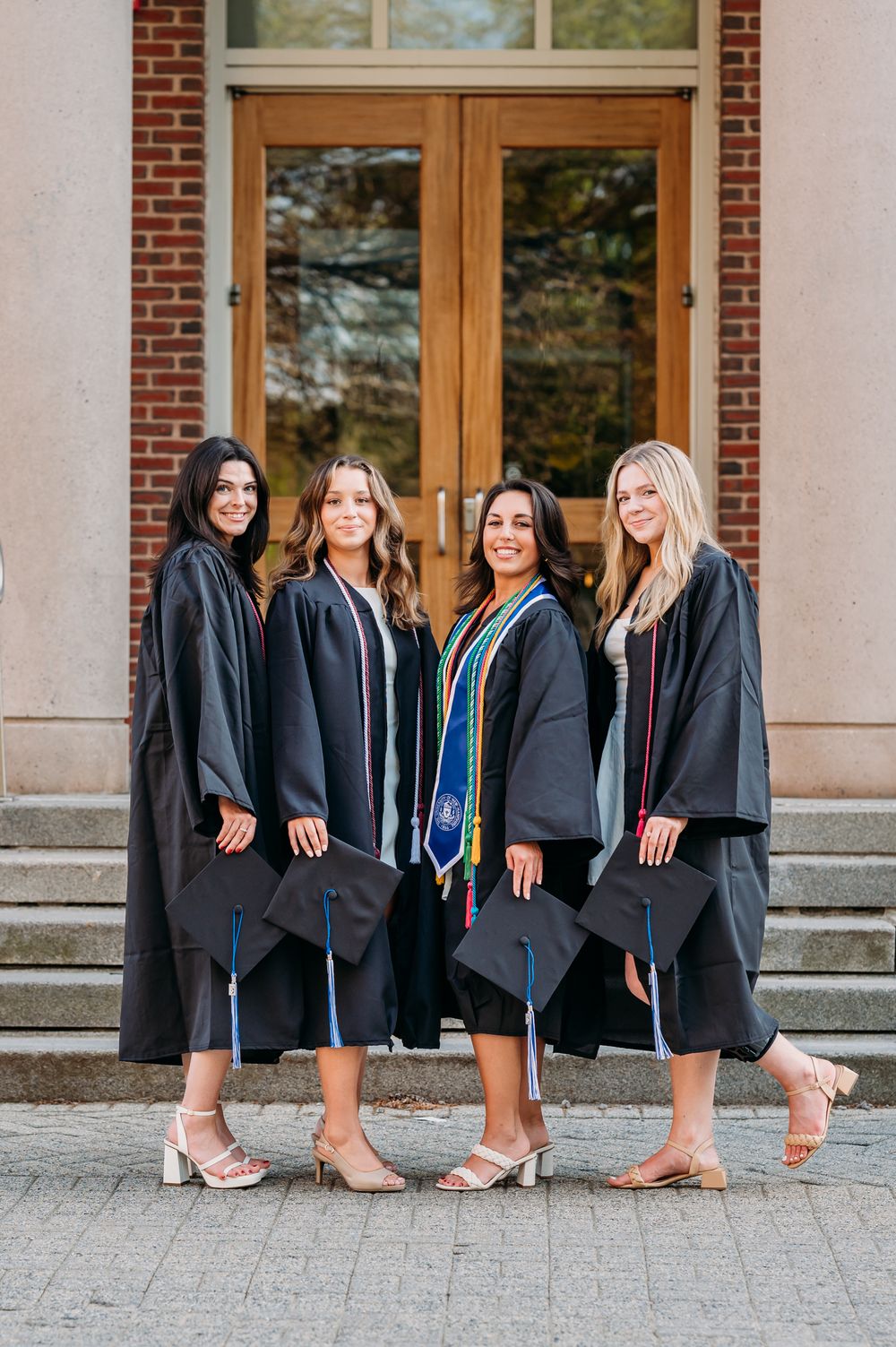 Group of graduates in black academic regalia and heels pose on stone steps in front of wooden doors at university building.