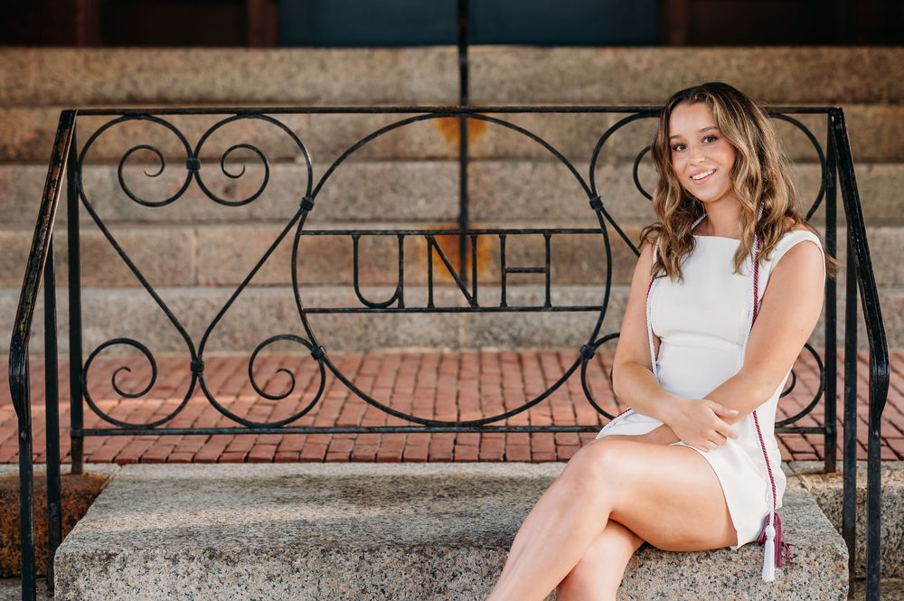 A person in a white dress sits on decorative metal steps with ornate scrollwork and UNH lettering.