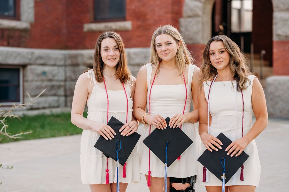 Three graduates in white dresses hold decorated mortarboard caps while posing together outside a brick building.