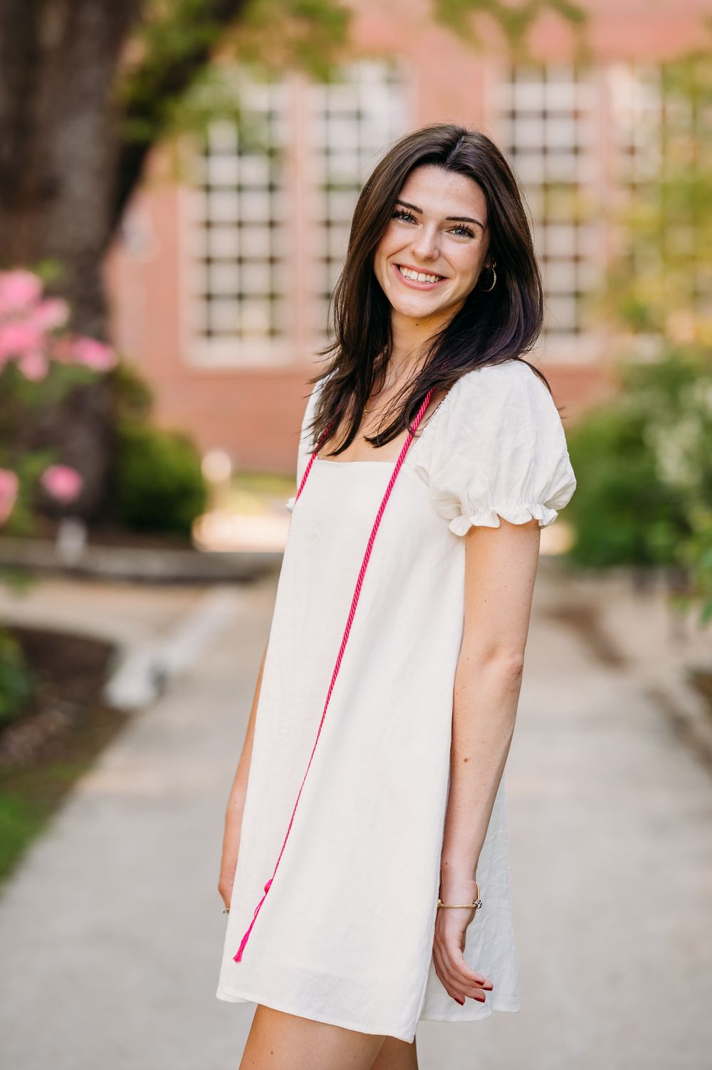 Young woman wearing a white summer dress poses outdoors on a sunny day with red brick building and flowers in background.