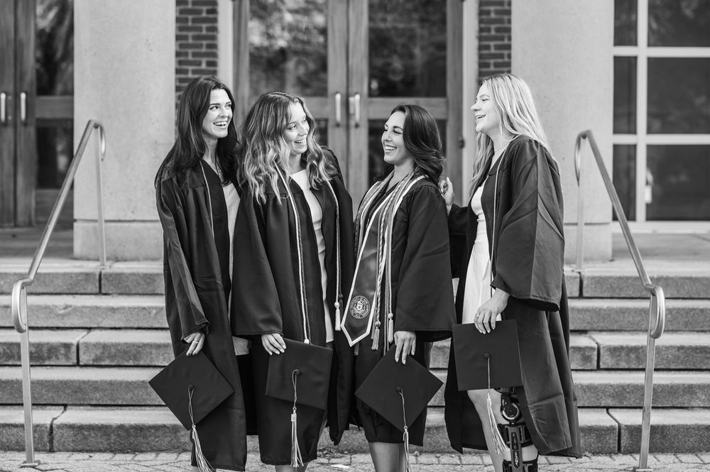 Four graduates wearing black gowns stand together on stone steps in front of a brick building in a black and white photo.
