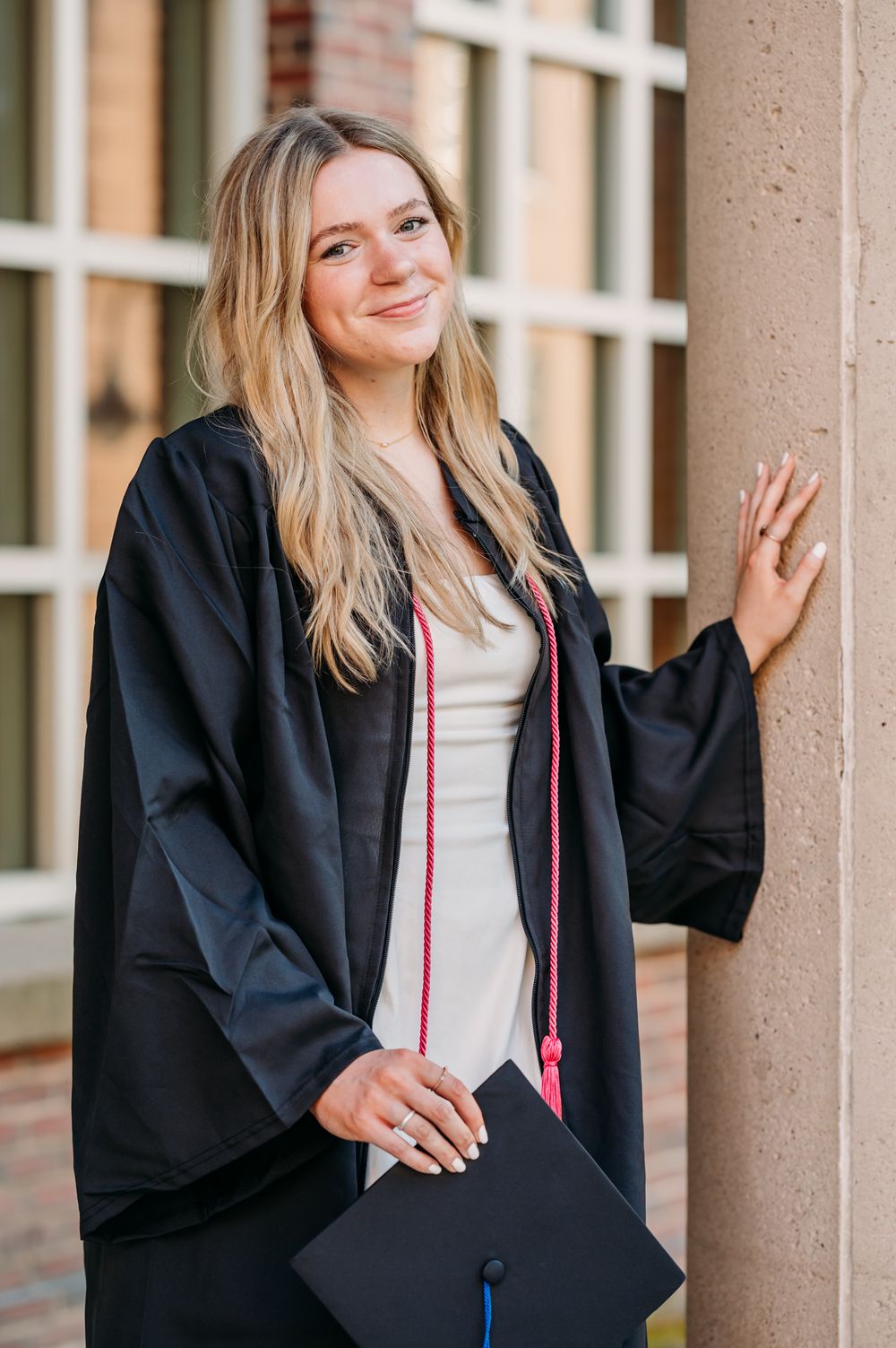 Graduate in black cap and gown holding diploma and smiling while posing against brick wall with columns.