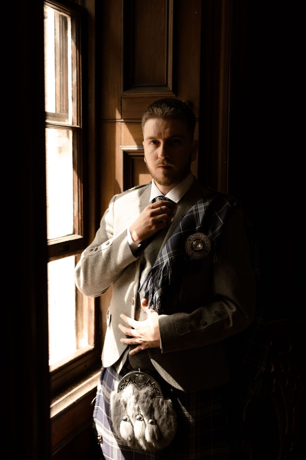 A figure in elegant formal attire adjusts their tie while standing in moody window lighting.