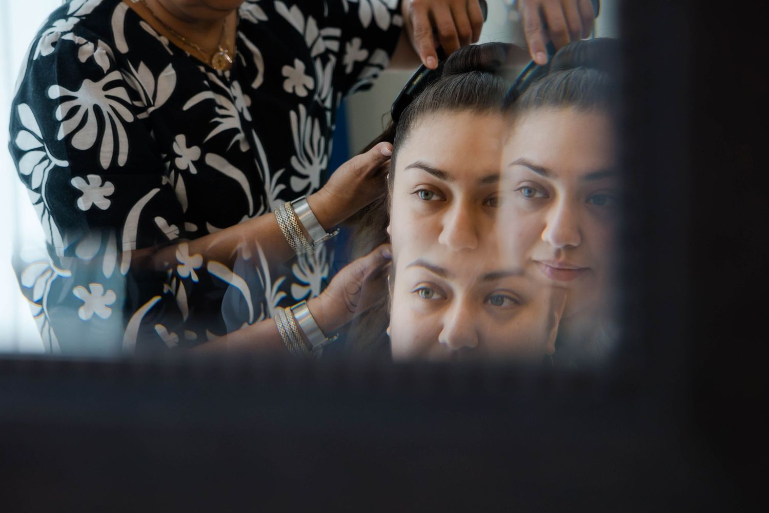 A reflected view through a mirror shows a person styling hair while wearing a black and white floral pattern shirt.