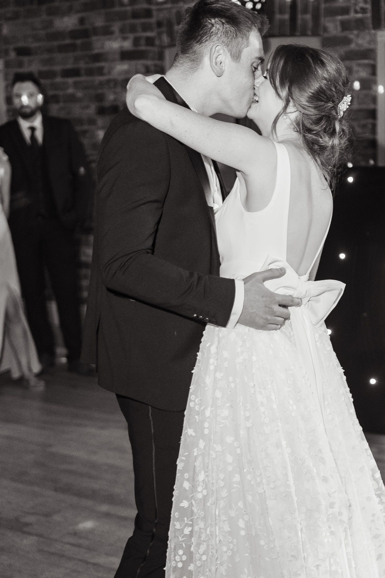 Couple shares intimate kiss during their first dance at wedding reception with brick wall backdrop and twinkling lights.