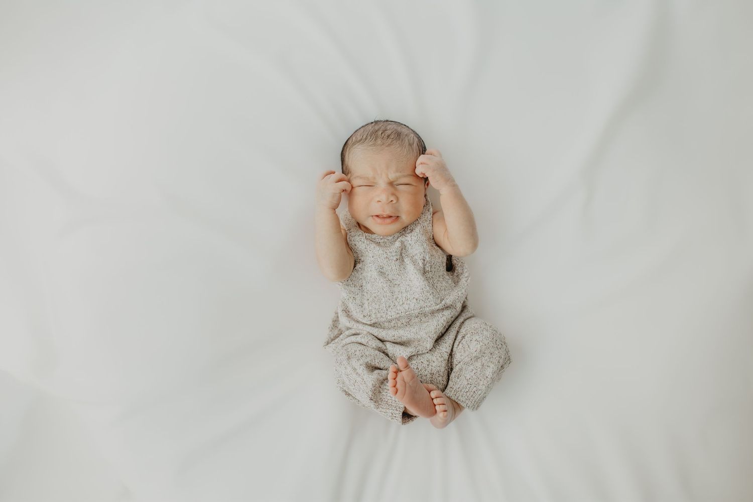 Newborn baby wearing gray outfit lies on white blanket while holding head with both hands.