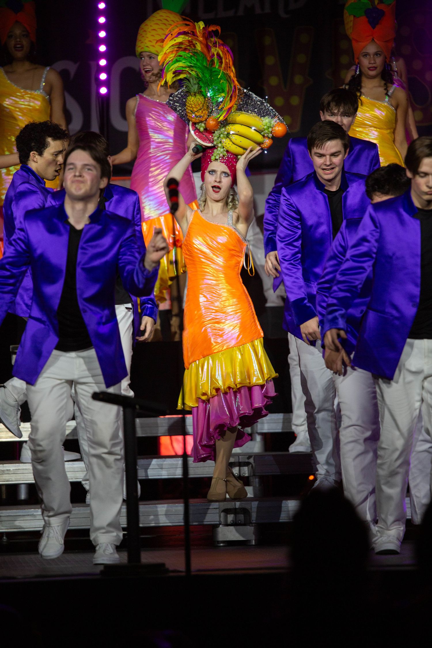 Performers in bright purple jackets and vibrant costumes dancing on stage during a colorful theatrical show.