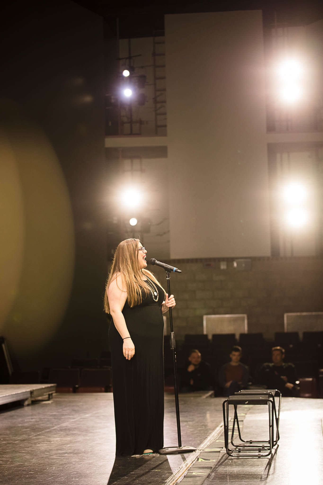 Solo performer in long black dress stands on stage under bright spotlights while singing into microphone during concert performance.