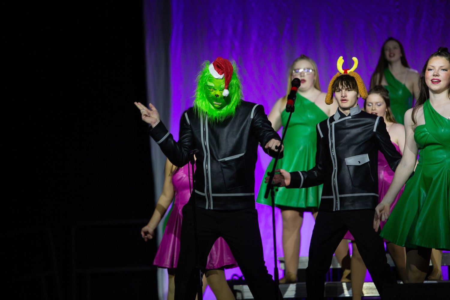 Performers in green dresses and black jackets dance on stage with dramatic purple lighting behind them.