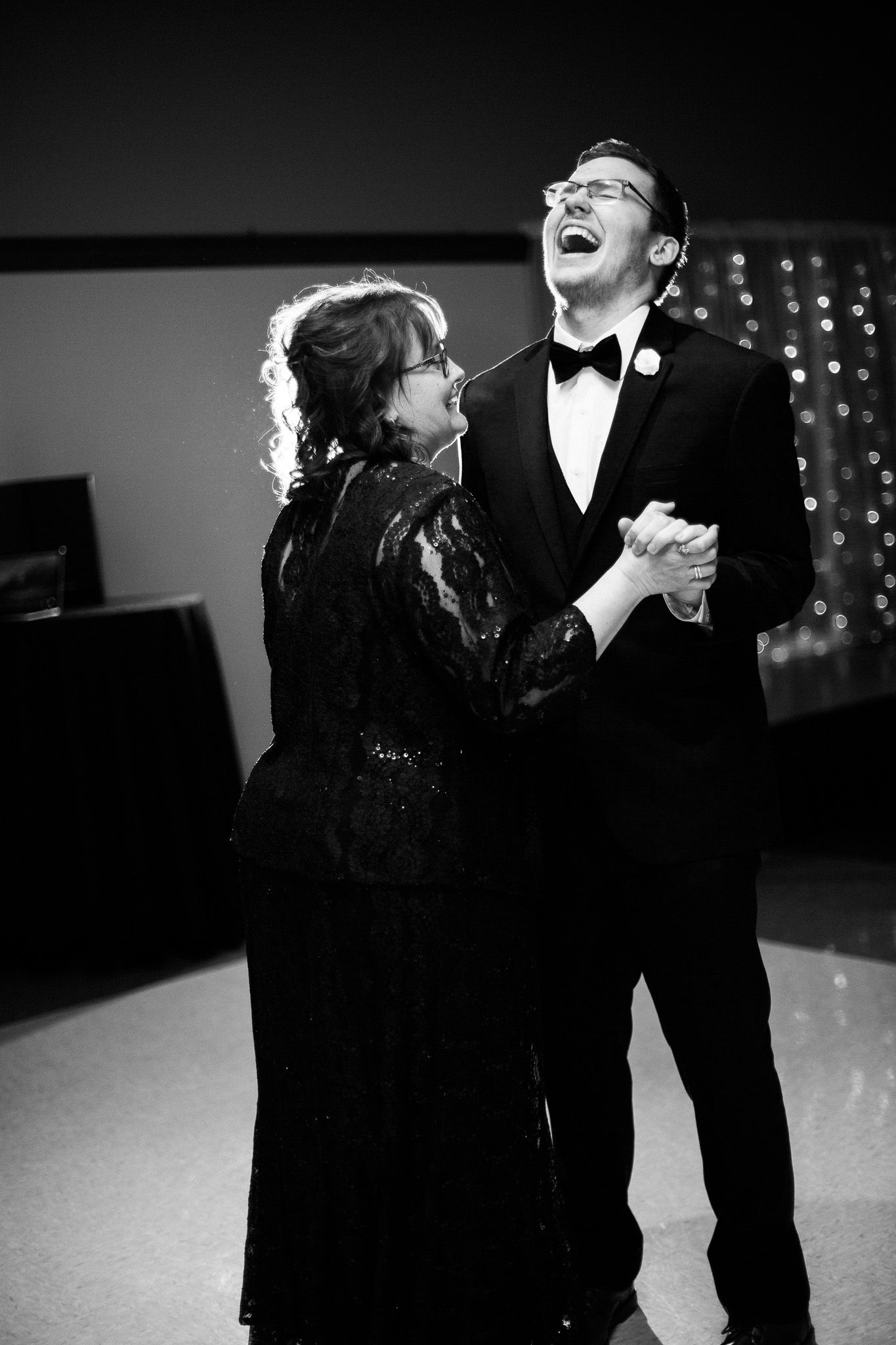 A couple shares a joyful dance moment in formal black attire at an indoor venue with twinkling lights in the background.