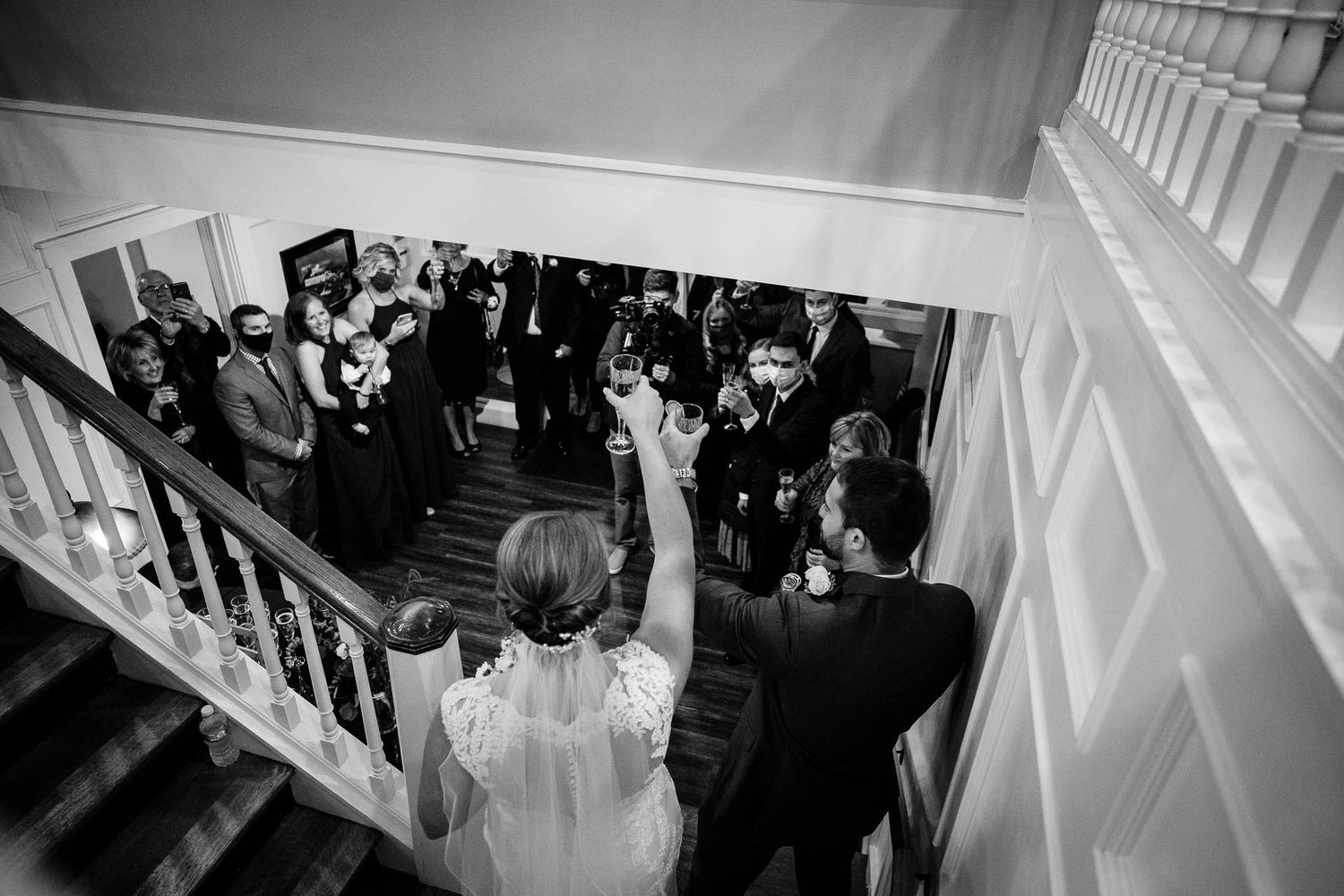 Black and white photo of wedding couple descending a grand staircase while guests cheer below at an elegant reception.