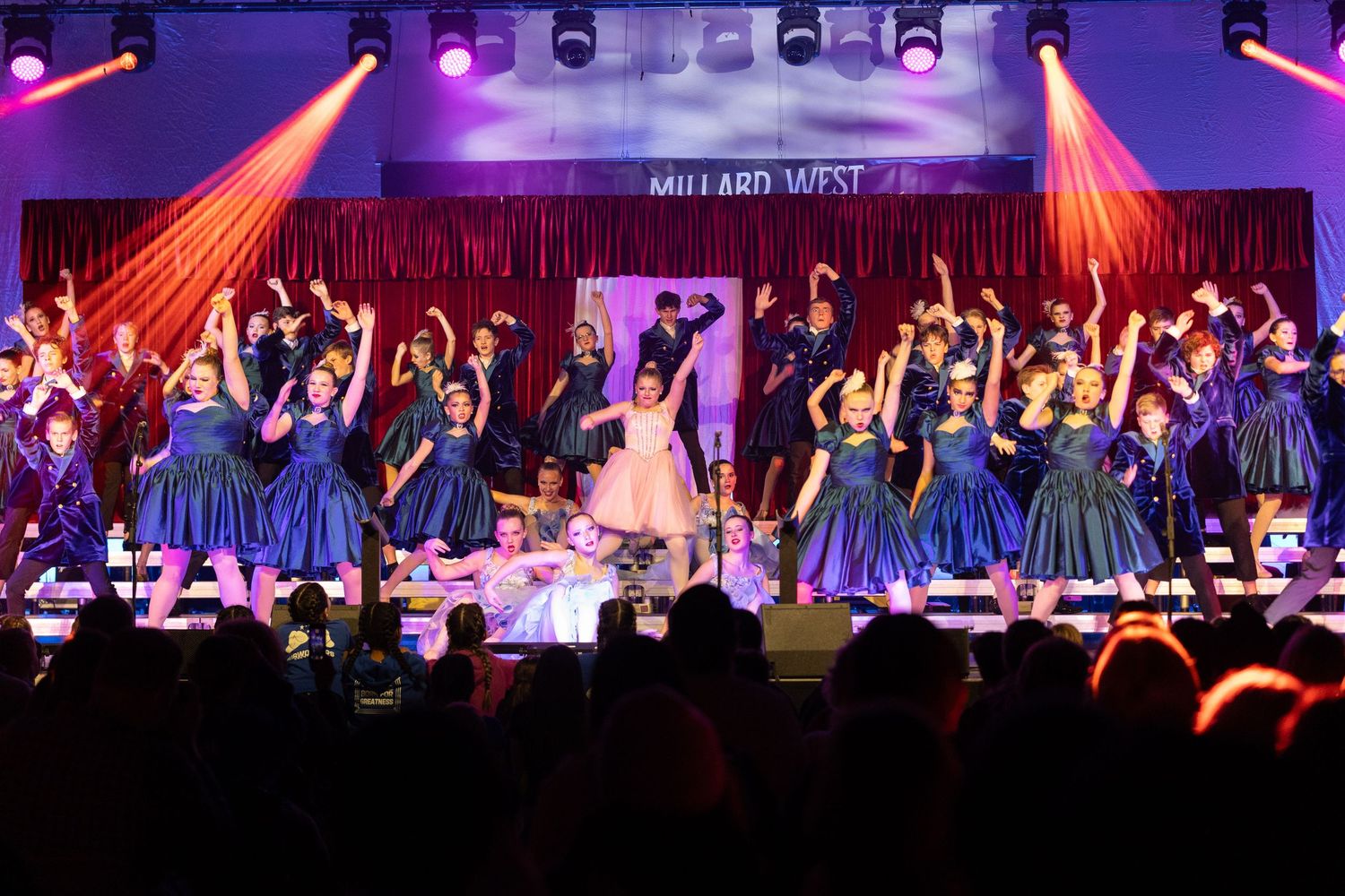 A large group of performers in blue dresses dance under dramatic red stage lighting at a theatrical production.