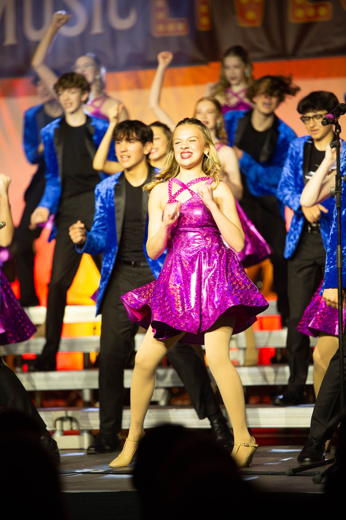 Group of performers in blue and pink sequined costumes dancing on stage during an energetic musical theater show.