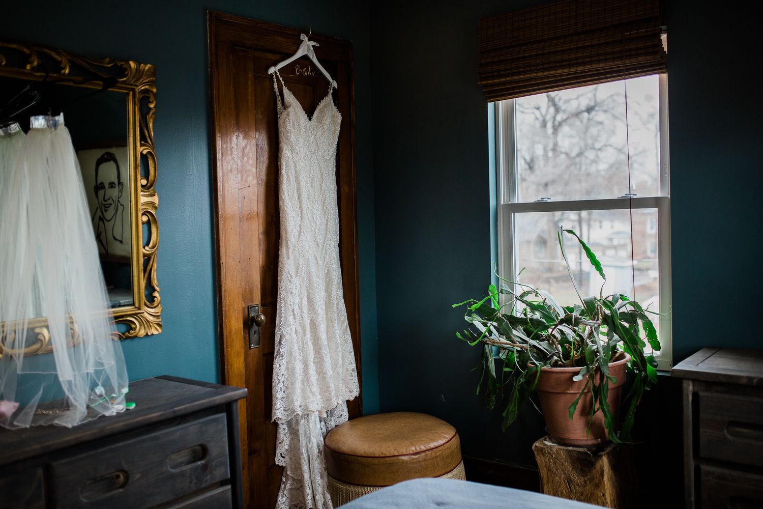 A white lace wedding dress hangs in a dark teal room with a window, dresser, and potted plant.