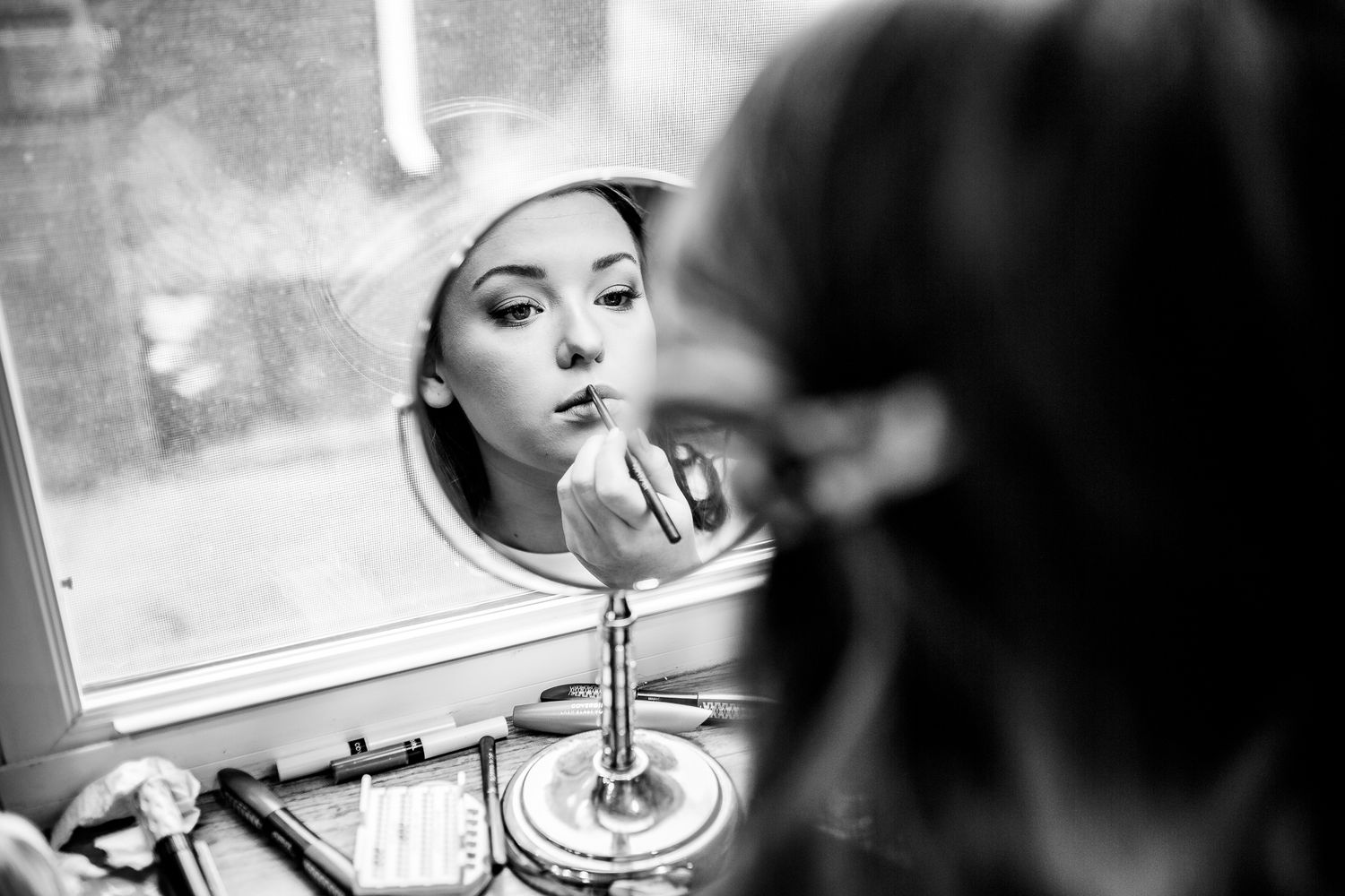 Black and white reflection of a person applying lipstick in a vanity mirror with dramatic lighting and composition.