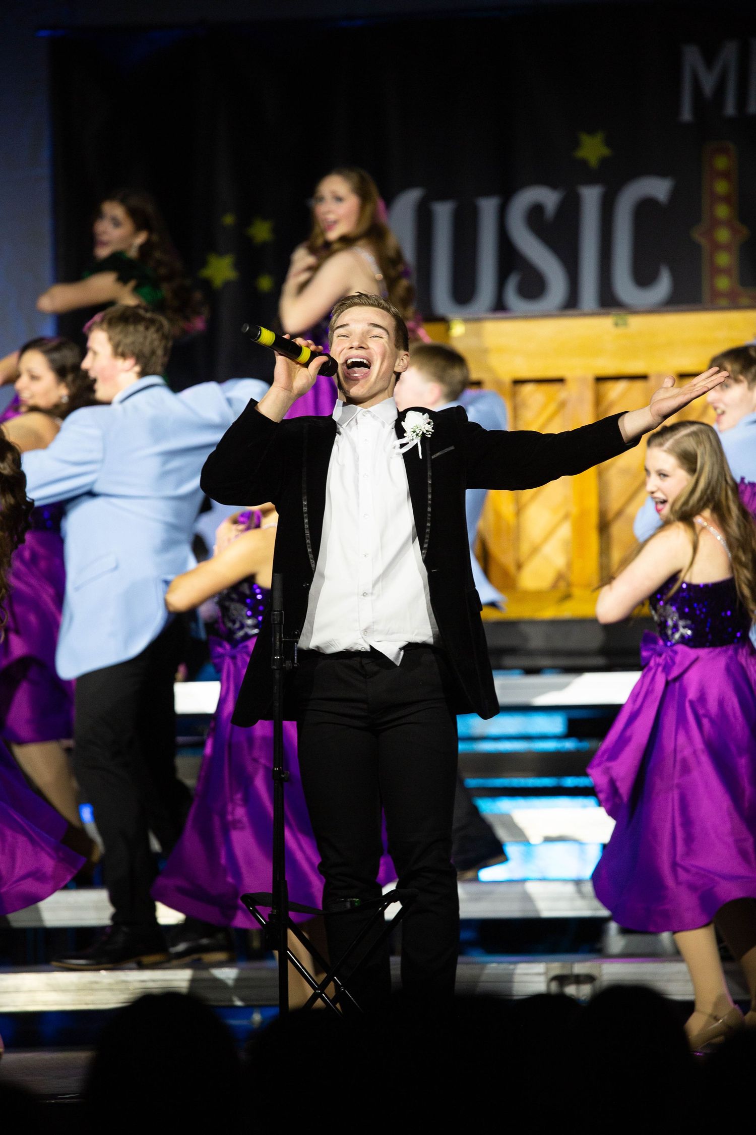 Performers in a show choir performance with singers in black tuxedos and purple dresses on risers with a MUSIC sign backdrop.