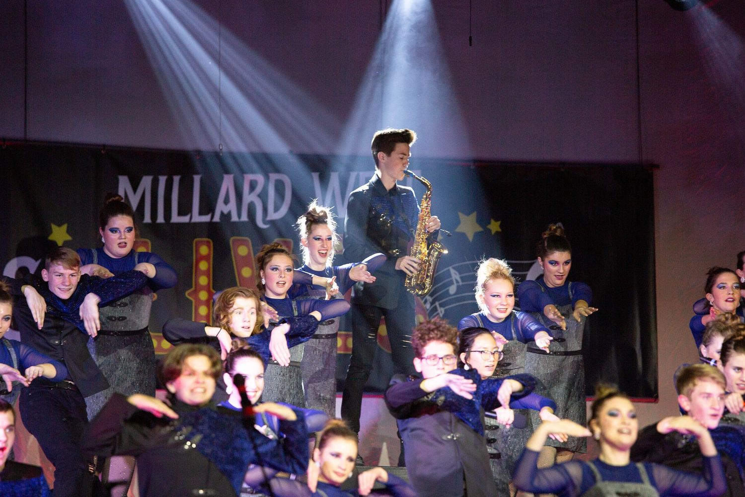 Students perform together on a darkened stage illuminated by spotlights during what appears to be a school production.