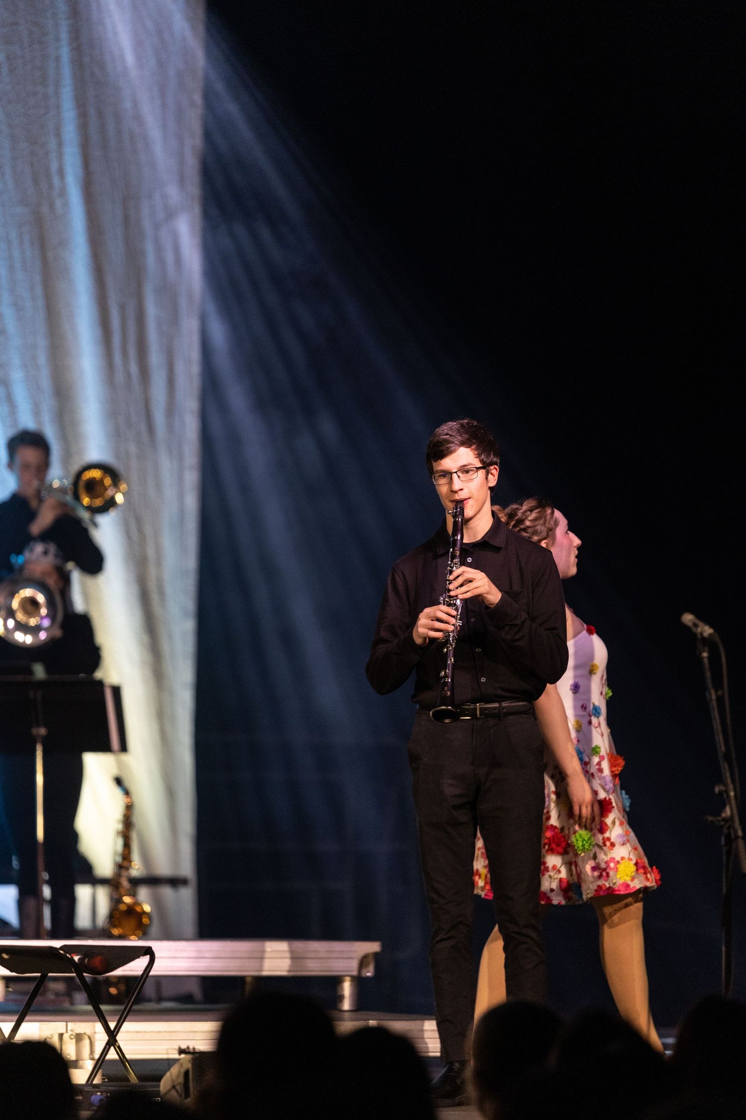 Musician in black attire performs on clarinet on dimly lit stage with band members and spotlights in background.