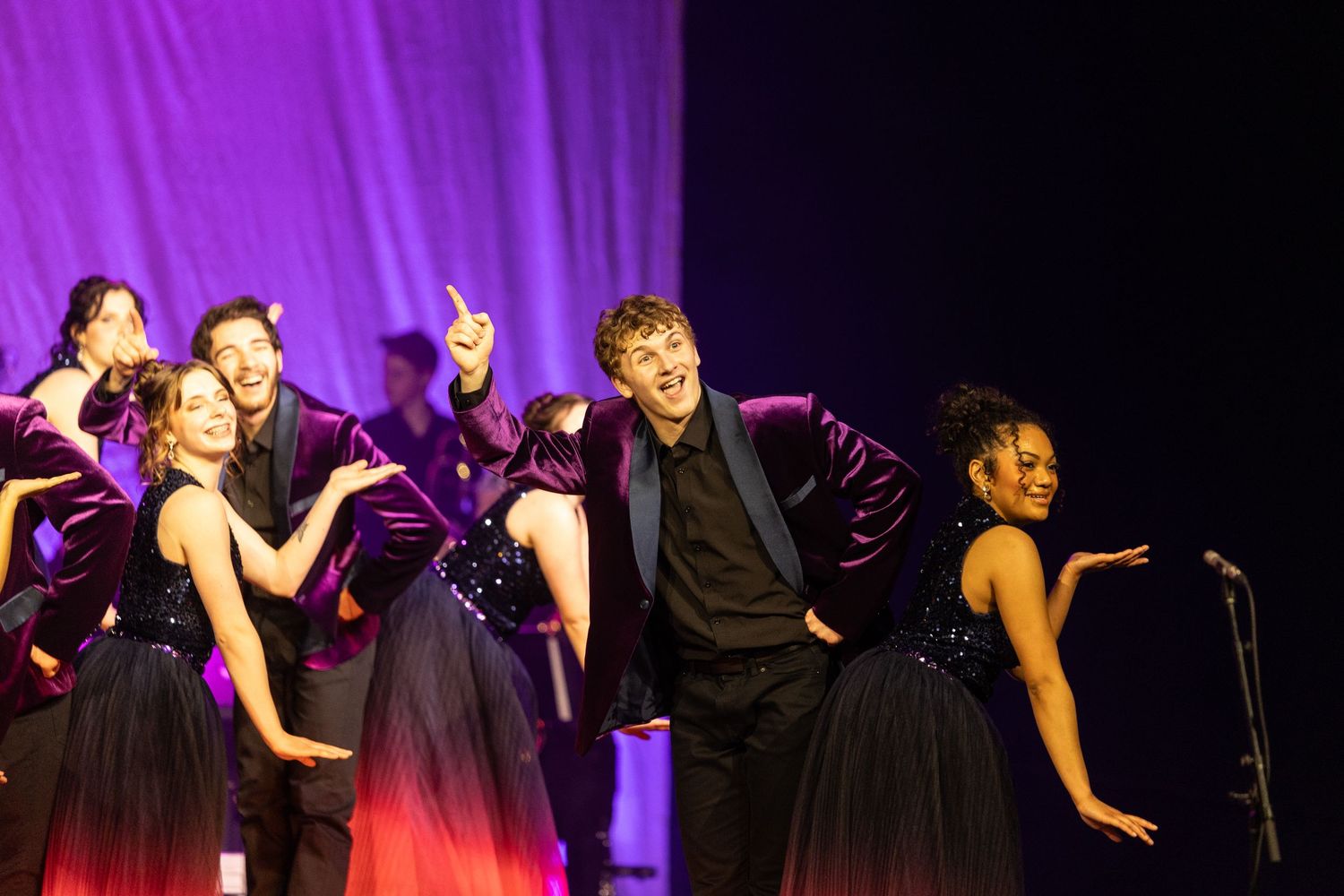 Performers in black dresses sing and dance energetically on a stage illuminated by purple and pink lighting effects.