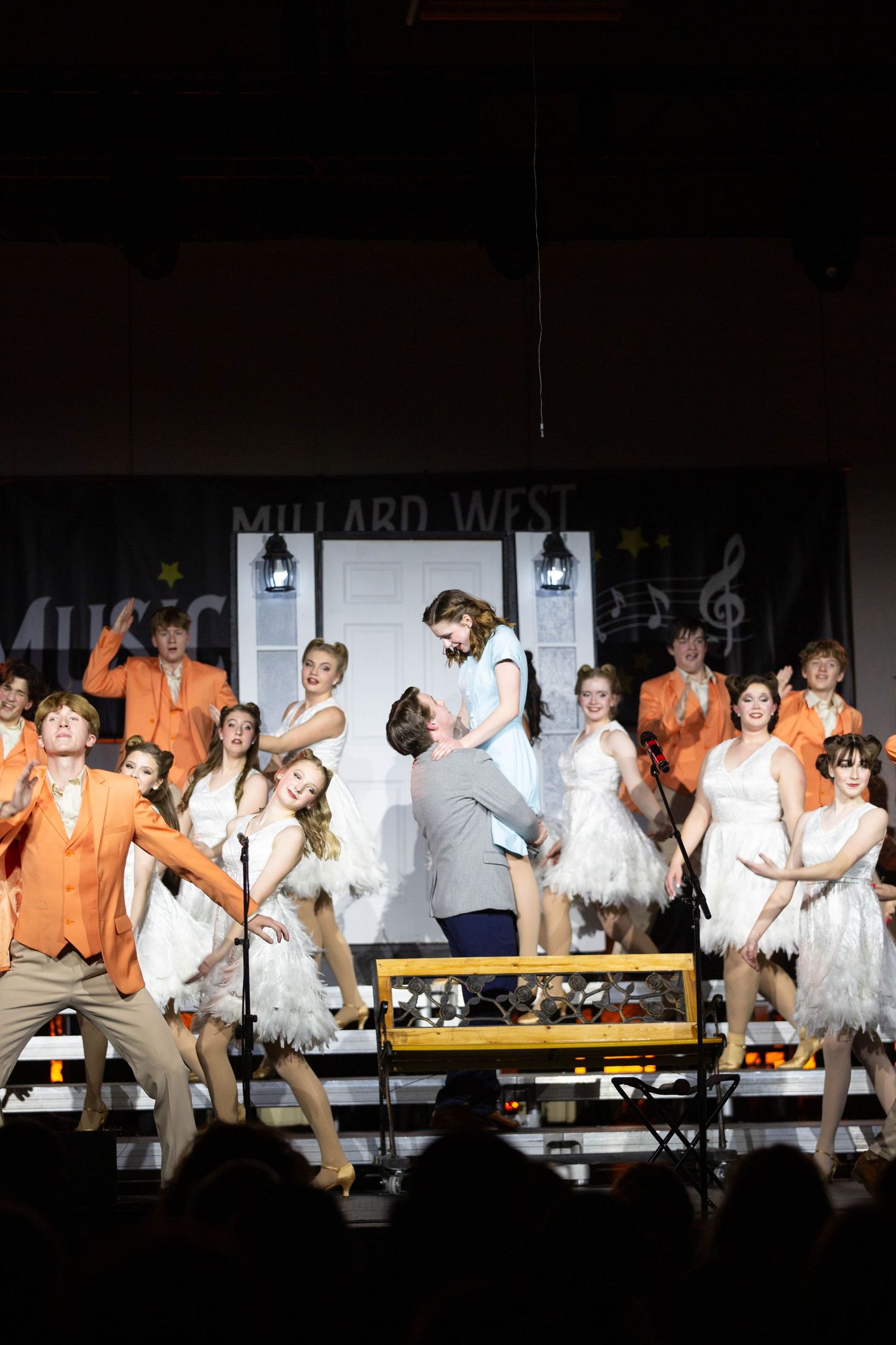 Performers in white and orange costumes dance energetically during a theatrical stage production number.