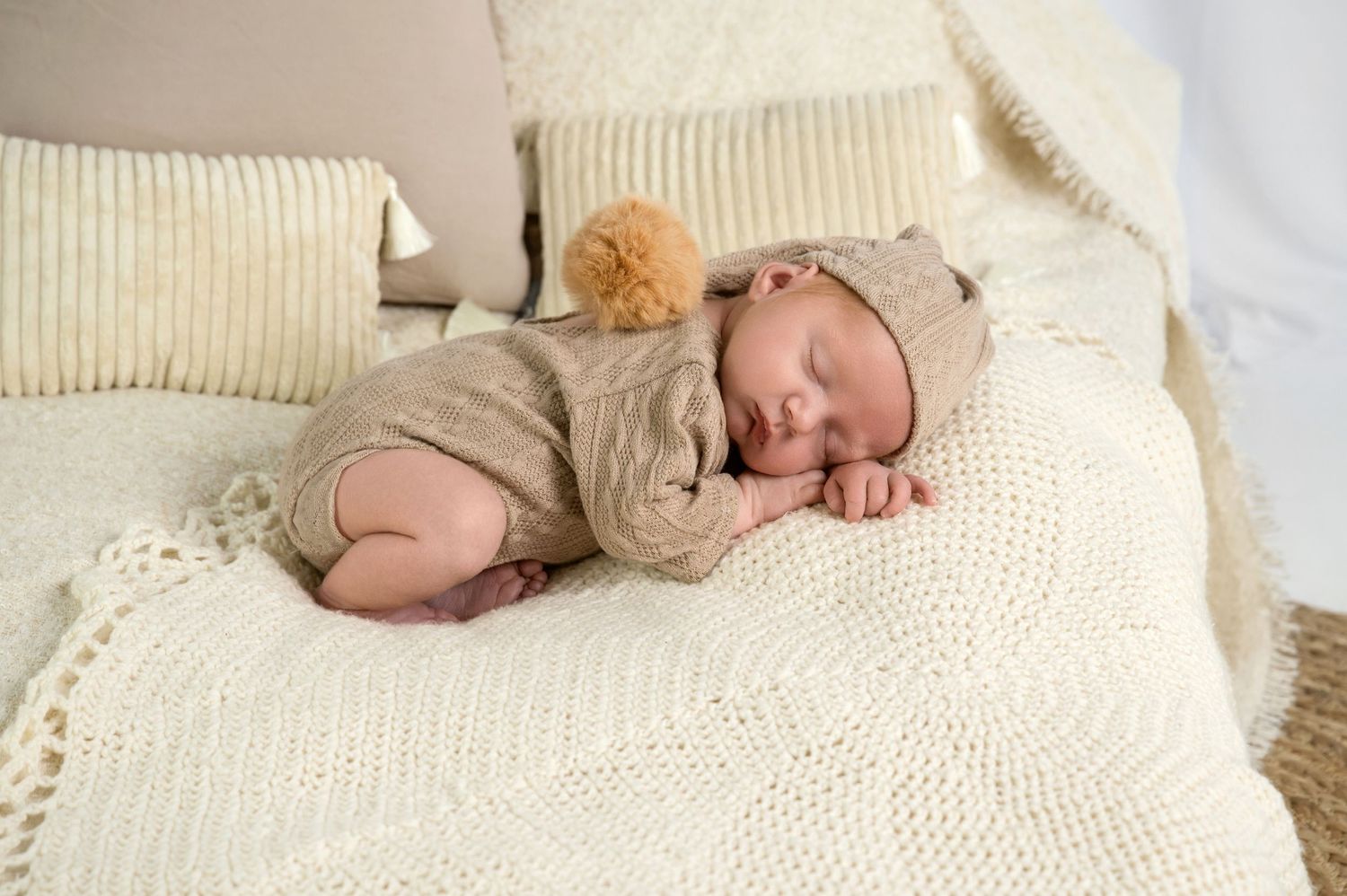 A sleeping baby in knit clothing rests peacefully on a cream-colored textured blanket.
