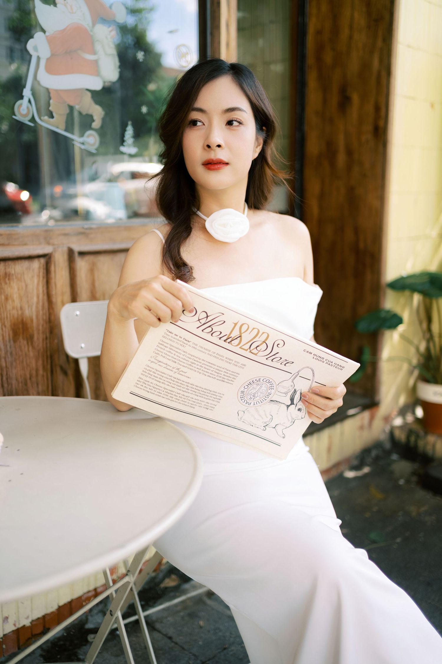 A person in an elegant white dress sits at a cafe table reading a newspaper.